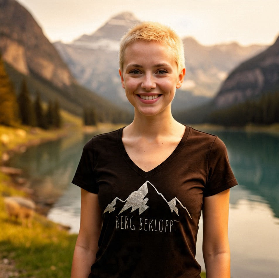 Eine junge blonde Frau, steht vor einem Bergsee und trägt das Berg bekloppt V-Neck Shirt